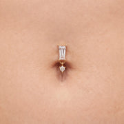 A close-up of a gold belly button ring with a baguette-cut clear stone and a dangling  clear stone, shown worn on skin.