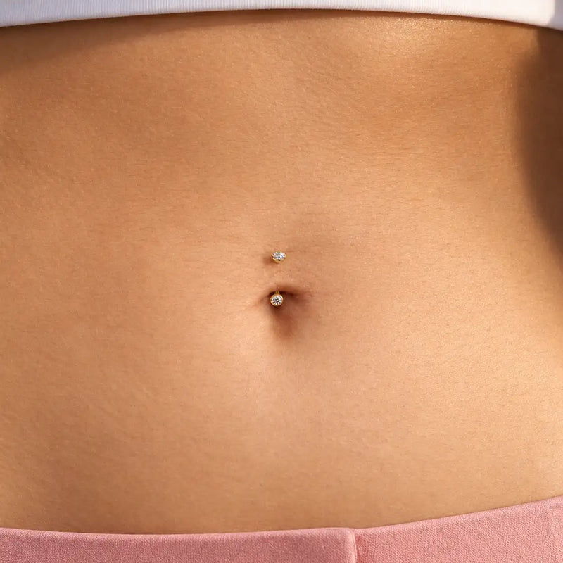 Close-up of a model's belly button piercing with a 3mm bezel-set curved barbell - two small clear stones set in 14k yellow gold
