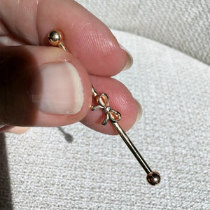14k gold bow industrial barbell held between fingers to show size, scale, and bow detail up close