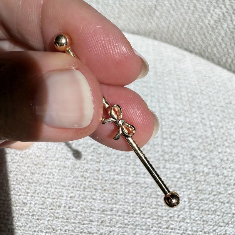 14k gold bow industrial barbell held between fingers to show size, scale, and bow detail up close