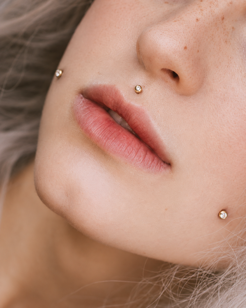 Cheek piercing with a gold bezel-set stud, set where a dimple would fall