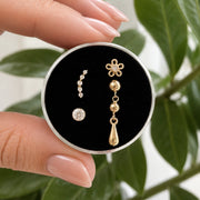 A close-up of a hand holding a round box with a black velvet insert, displaying a pair of gold dangle earrings and a small round stud earring. The background features soft-focus green leaves.