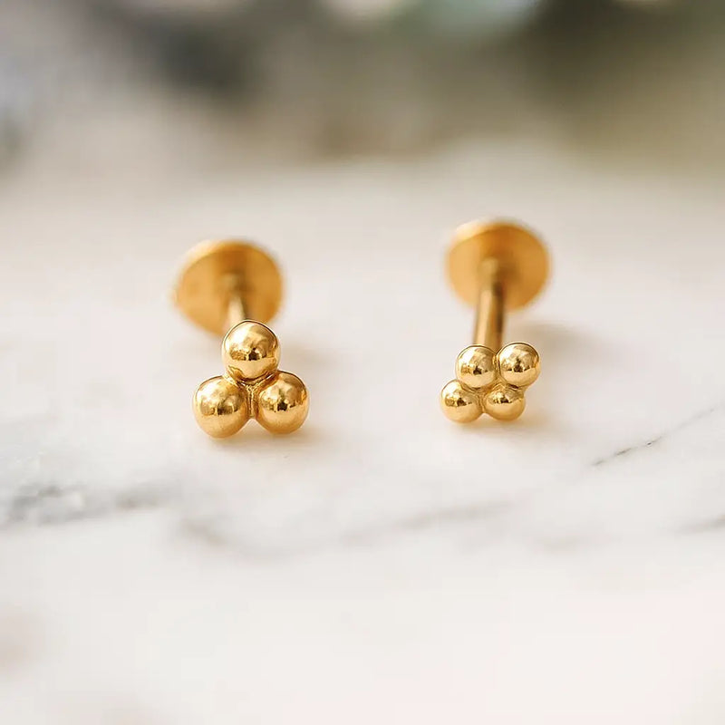 Pair of 14k yellow gold tri bead cluster threadless studs resting on marble surface, front and angled views