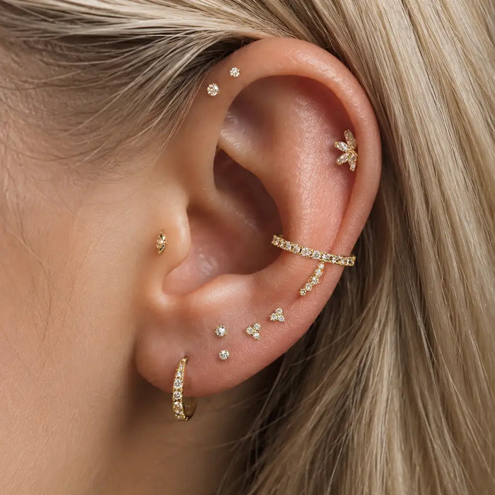 A close-up of a model's ear adorned with multiple gold ear piercings, including a curved ear cuff, a small hoop, several stud earrings, and a flower-shaped cartilage piercing, against a background of blonde hair.