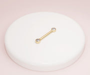 14K yellow gold CZ bezel nipple barbell resting on white ceramic dish against pink backdrop