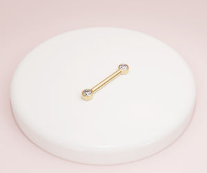 14K yellow gold CZ bezel nipple barbell resting on white ceramic dish against pink backdrop