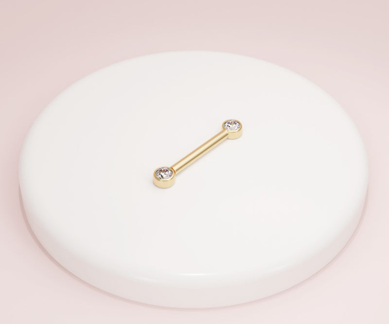 14K yellow gold CZ bezel nipple barbell resting on white ceramic dish against pink backdrop