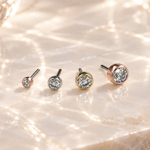 Four round bezel threadless ends with clear gemstones in white gold, yellow gold, and rose gold settings, arranged in ascending size order on a light beige marble surface.
