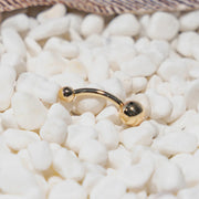 14k yellow gold double ball belly ring resting on white pebbles, close-up lifestyle detail shot