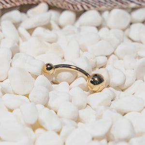 14k yellow gold double ball belly ring resting on white pebbles, close-up lifestyle detail shot