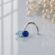 Aquamarine and blue sapphire white gold nose ring on a reflective glass with flowers in the background.