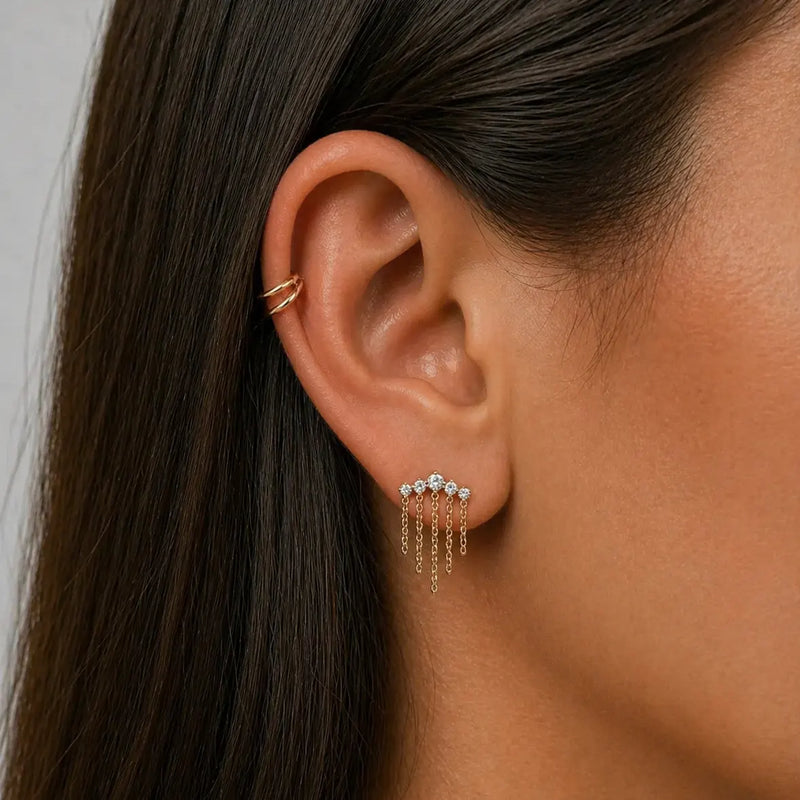 Close-up of an ear wearing a gold earring with small stones and dangling chains with a gold double hoop helix piercing.