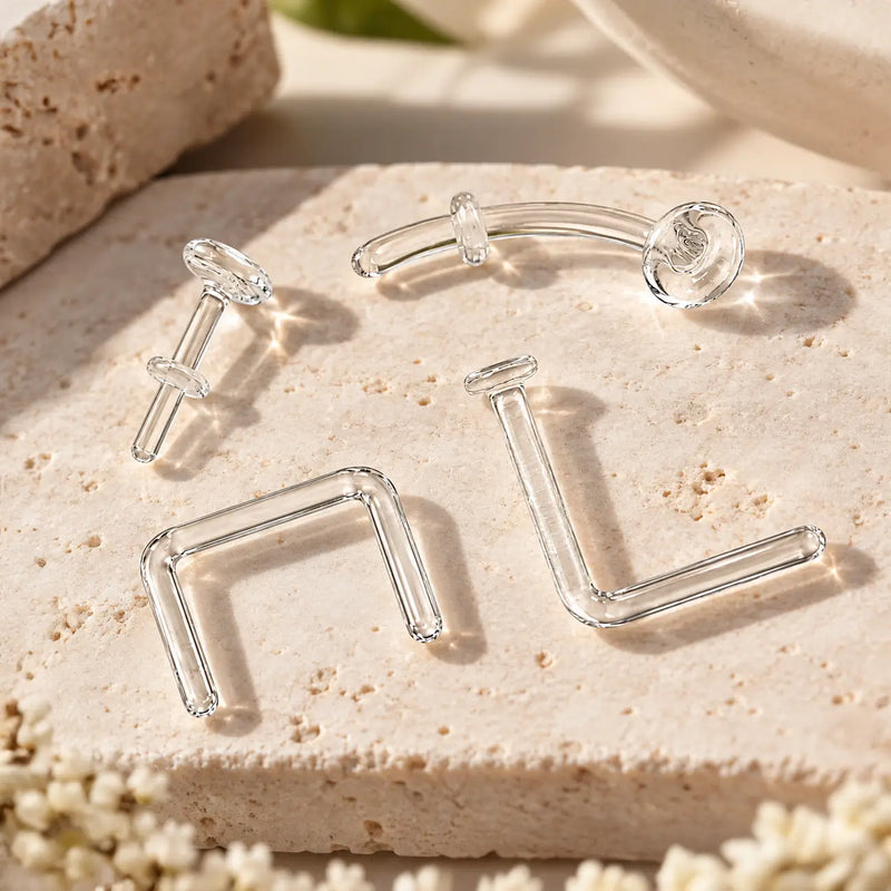 Four clear glass piercing retainers on travertine - flat back labret, curved barbell, septum staple and nose L-bend styles