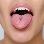 Model wearing a 14k gold heart tongue ring with a pink heart-cut gemstone, tongue extended, front view