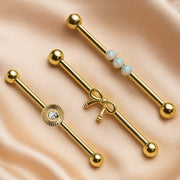 Bow industrial barbell in 14k gold displayed alongside sunburst CZ and opal style barbells on satin
