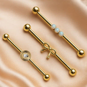 Bow industrial barbell in 14k gold displayed alongside sunburst CZ and opal style barbells on satin