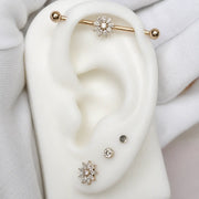 A close-up of a white ear display form wearing multiple ear piercings: an industrial barbell with a flower-shape, two small studs at the lobe, and a larger flower-shaped stud at the lobe.
