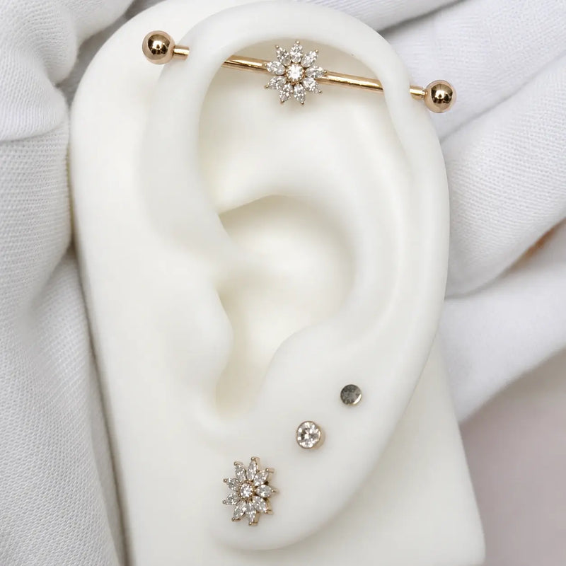A close-up of a white ear display form wearing multiple ear piercings: an industrial barbell with a flower-shape, two small studs at the lobe, and a larger flower-shaped stud at the lobe.