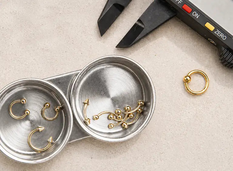 Gold piercing jewelry in a metal dish with calipers for jewelry manufacturing