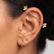 A close-up of a person's ear showcasing the Golden Lotus industrial barbell, along with other small hoop and stud earrings.