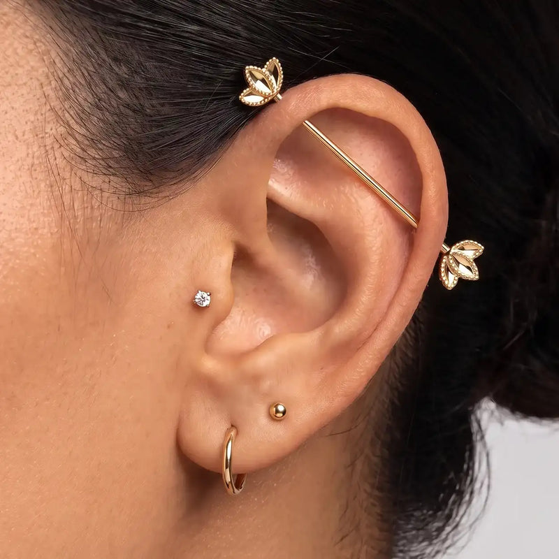 A close-up of a person's ear showcasing the Golden Lotus industrial barbell, along with other small hoop and stud earrings.