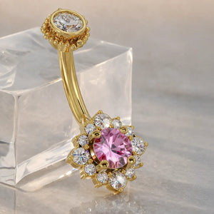 14k yellow gold halo belly ring with pink sapphire center and diamond halo, angled on crystal block