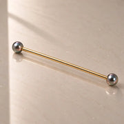 A gold industrial barbell with peacock pearl ends, resting diagonally on a light beige reflective surface.