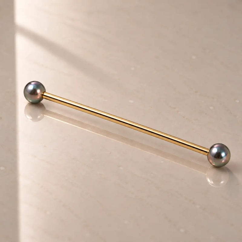 A gold industrial barbell with peacock pearl ends, resting diagonally on a light beige reflective surface.