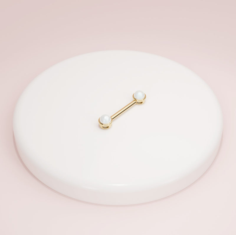 14K yellow gold pearl nipple barbell resting on white ceramic dish against pink background