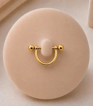A yellow gold stirrup nipple piercing with a displayed on a round beige jewelry stand.