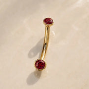 A curved gold barbell with two round faceted ruby gemstones, displayed on a light beige marble surface.