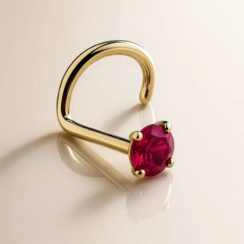 A 14k gold ruby gemstone nose ring in a four-prong setting, displayed on a clean, light beige reflective surface.