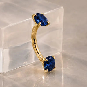 Gold curved barbell piercing jewelry with two round sapphire gemstones, displayed on a clear acrylic block over a reflective surface.
