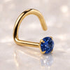 A yellow gold nose ring twist with a blue sapphire, shown on a reflective light marble surface.