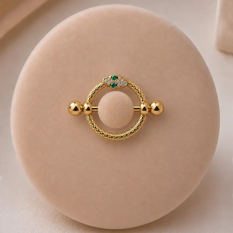 A yellow gold snake nipple shield, set with small green and clear stones, displayed on a velvet stand.