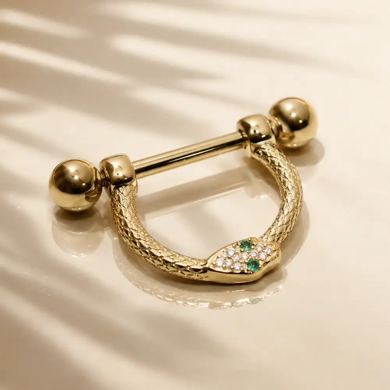 14k yellow gold nipple stirrup with a textured snake-head design with diamonds and emeralds, photographed on a warm cream surface with soft shadow overlays.