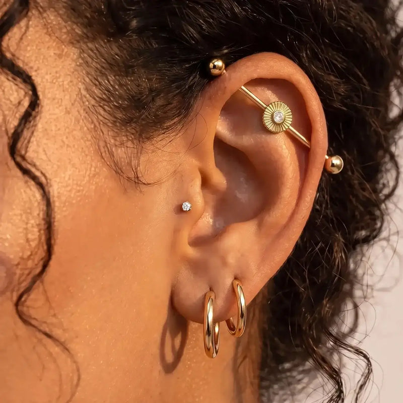 Close-up of a person's ear adorned with multiple gold ear piercings, including hoop earrings, small stud and the Soleil industrial barbell.