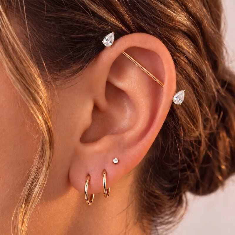 A close-up of a person's ear showcasing a multi-piece gold hoop set, including an industrial barbell with teardrop gemstone ends.