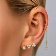 A close-up of a model's ear showcasing a multi-piece gold ear cuff set with clear gemstones, including a small gold hoop, a chain with a dangling heart, a square stud, and a cluster of round diamonds.