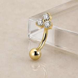 A gold curved barbell piercing with three round diamonds at the top, displayed leaning on a clear acrylic stand over a light beige reflective surface.