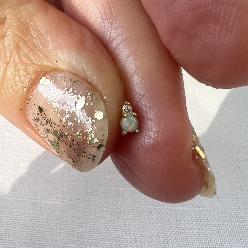 Tiny 14k gold opal and diamond piercing stud end pinched between fingertips, close-up scale view