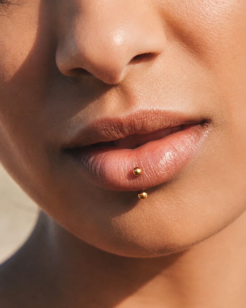 Vertical labret piercing with a gold curved barbell, both ball ends visible on and below the lower lip