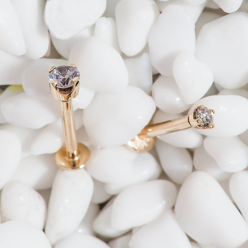 Two 14k rose gold 3mm CZ flat back labret studs nestled among white pebbles, lifestyle close-up