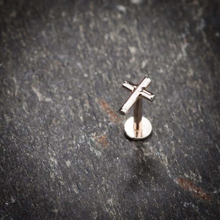 14K rose gold cross labret stud standing upright on slate stone surface for scale