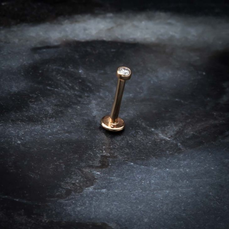 14k rose gold bezel-set diamond flat back labret standing upright on dark stone surface, lifestyle shot