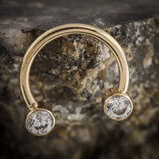 14k yellow gold circular barbell with clear CZ bezel ends resting against rough stone surface