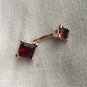 14k rose gold belly ring with red princess-cut CZ stones laid flat on linen fabric