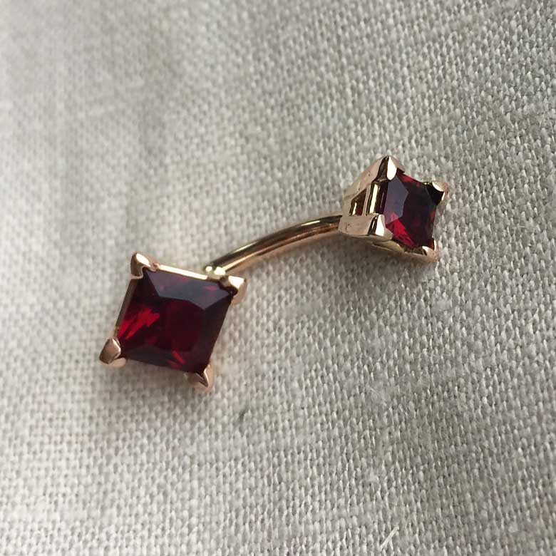 14k rose gold belly ring with red princess-cut CZ stones laid flat on linen fabric