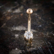 14k rose gold navel ring with round CZ prong setting, upright on dark stone surface