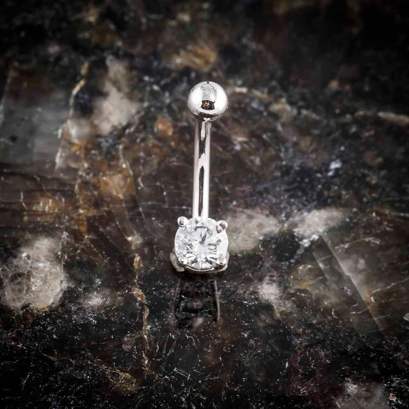 14k white gold CZ navel ring standing upright on dark mineral surface, full-length editorial view